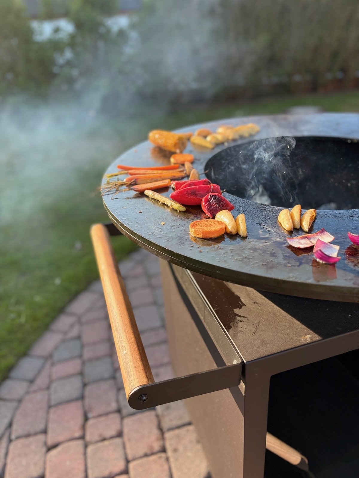 Gemüse brutzelt auf dem OFYR-Grill, dahinter der Garten